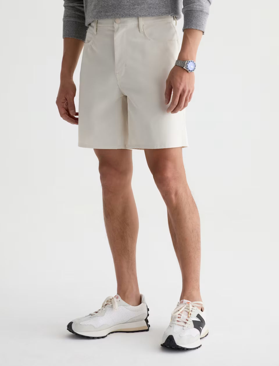 A person with an athletic build stands against a plain background, wearing a gray sweater, AG Jeans’ off-white Bay Short, white and black sneakers, and a silver wristwatch.