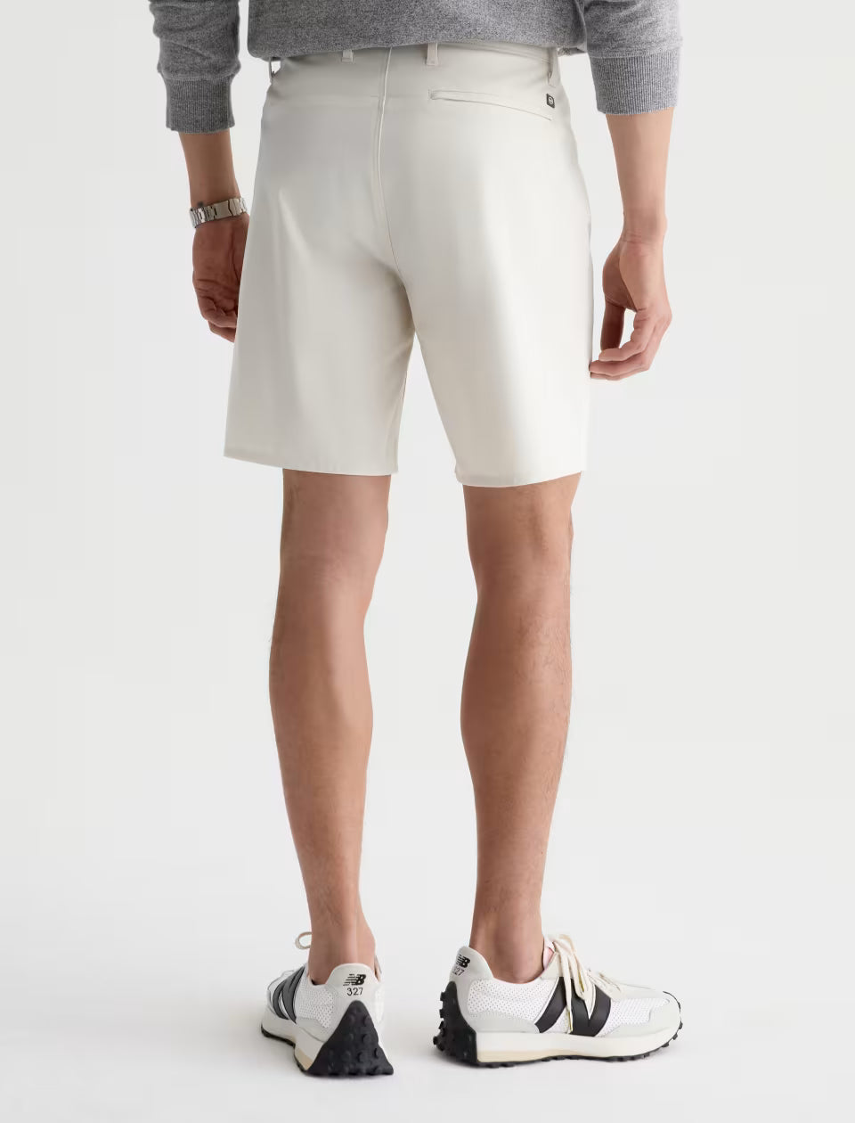 An athletic model is shown from the back wearing a gray sweater, AG Jeans Bay Short in a modern off-white summer silhouette that reaches the knees, and white sneakers with black accents.