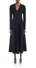 A person wears the Maria McManus Button Front Dress—a long-sleeved, black button-front dress with corozo nut buttons and a flared skirt—paired with white shoes, standing against a plain white background.