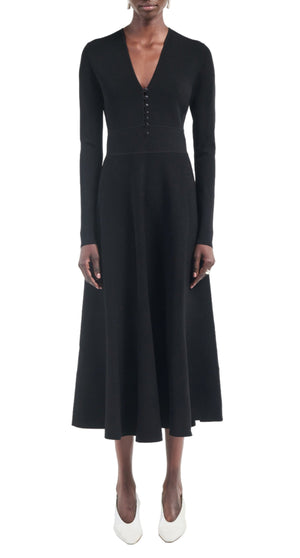 A person wears the Maria McManus Button Front Dress—a long-sleeved, black button-front dress with corozo nut buttons and a flared skirt—paired with white shoes, standing against a plain white background.