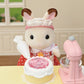 A delightful scene from the Calico Critters Village Cake Shop Starter Set features a toy bunny in a red dress decorating a cake with pink icing using a piping bag. Surrounded by cake decorations, with a pink mixer and mixing bowl on the table, this charming set creates the perfect atmosphere for imaginative play in your very own cake shop.