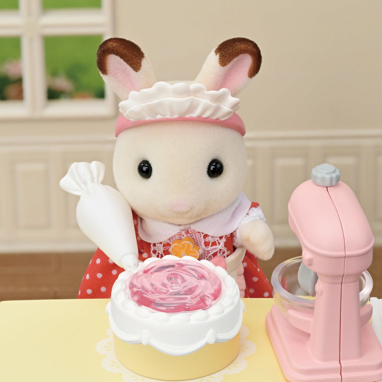 A delightful scene from the Calico Critters Village Cake Shop Starter Set features a toy bunny in a red dress decorating a cake with pink icing using a piping bag. Surrounded by cake decorations, with a pink mixer and mixing bowl on the table, this charming set creates the perfect atmosphere for imaginative play in your very own cake shop.
