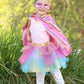 A young child wearing the Great Pretenders Super-duper Tutu, Cape & Mask (4-6Y) set stands outdoors on grass with green plants in the background—the perfect moment for dress-up adventures.