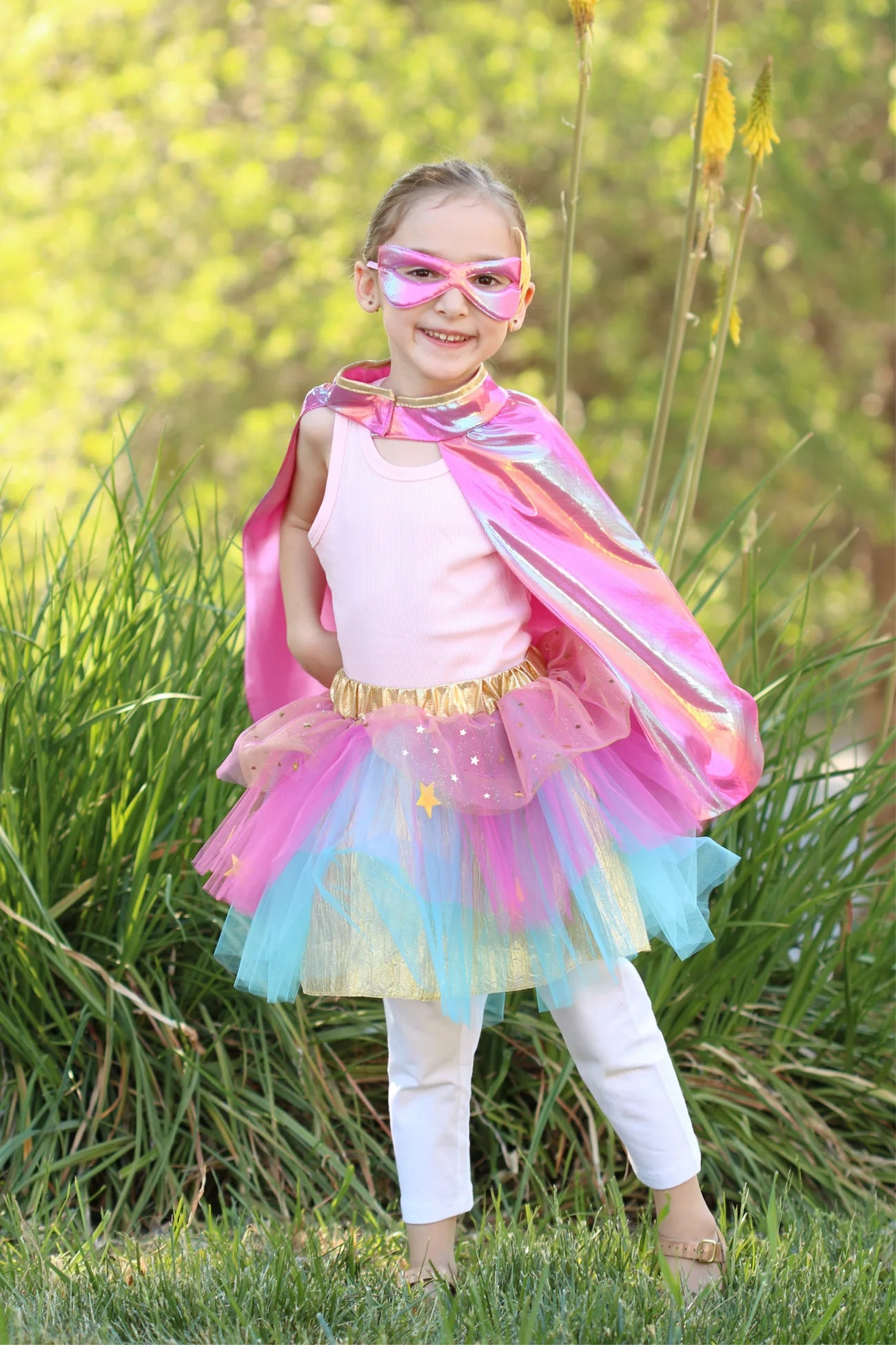 A young child wearing the Great Pretenders Super-duper Tutu, Cape & Mask (4-6Y) set stands outdoors on grass with green plants in the background—the perfect moment for dress-up adventures.