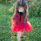 Wearing the Great Pretenders Glitter Ladybug Set, a young child faces away from the camera on a grassy lawn, showing off delicate ladybug wings and a playful tutu skirt in this charming outdoor scene.