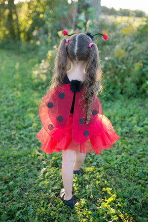 Wearing the Great Pretenders Glitter Ladybug Set, a young child faces away from the camera on a grassy lawn, showing off delicate ladybug wings and a playful tutu skirt in this charming outdoor scene.