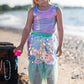 A young child wears the Great Pretenders Bling Bling Mermaid Skirt & Hairclip Set, featuring a pink tulle sequin skirt, and stands on a sandy beach near a wooden chest with a bubble wand and the ocean in the background.