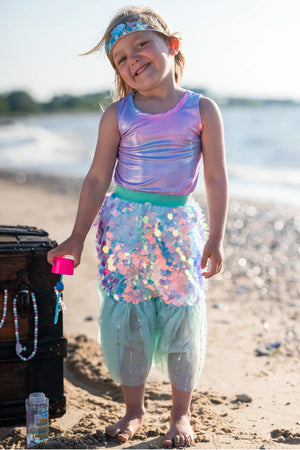 A young child wears the Great Pretenders Bling Bling Mermaid Skirt & Hairclip Set, featuring a pink tulle sequin skirt, and stands on a sandy beach near a wooden chest with a bubble wand and the ocean in the background.