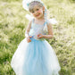 A young child stands outdoors on grass, smiling at the camera while wearing the Great Pretenders Sequins Princess Dress featuring a blue sparkly bodice, tulle skirt, and a matching blue hair braid accented with a flower headband.