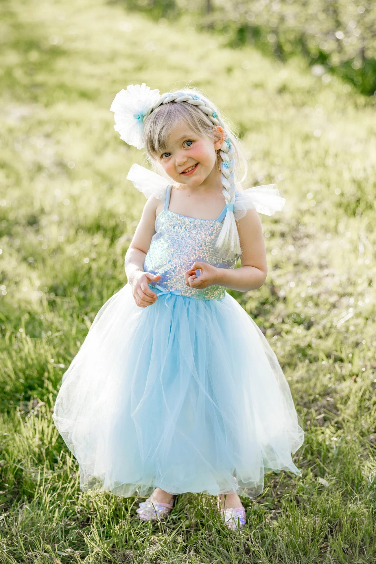 A young child stands outdoors on grass, smiling at the camera while wearing the Great Pretenders Sequins Princess Dress featuring a blue sparkly bodice, tulle skirt, and a matching blue hair braid accented with a flower headband.