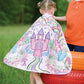 A child wearing the Great Pretenders Princess Color in Cape (for ages 4-7), featuring a colorful castle and unicorn design, stands on painted tires outdoors beside another child in an orange shirt.