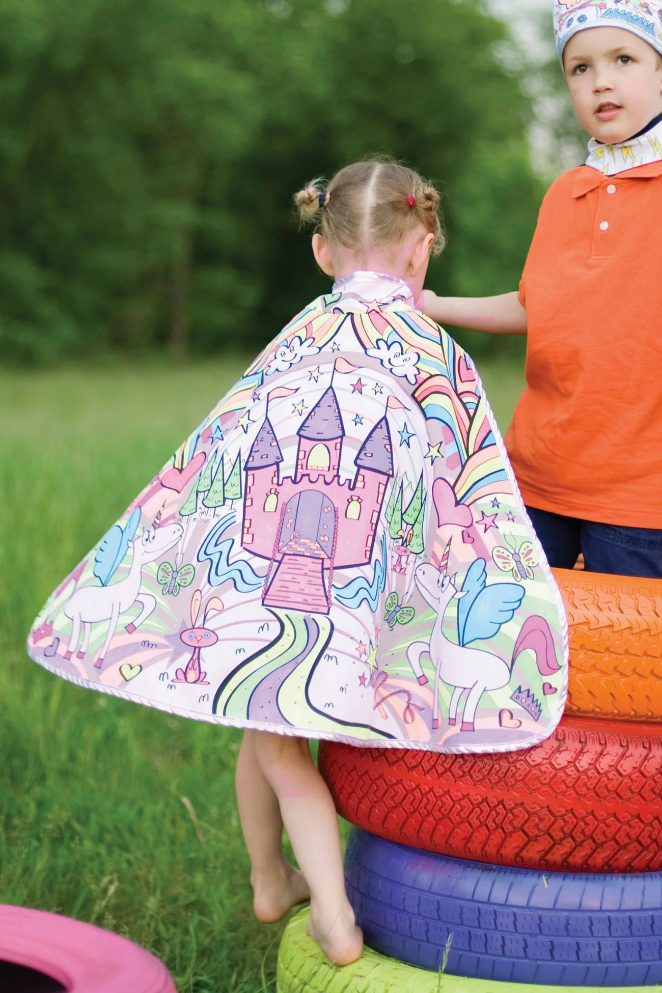 A child wearing the Great Pretenders Princess Color in Cape (for ages 4-7), featuring a colorful castle and unicorn design, stands on painted tires outdoors beside another child in an orange shirt.