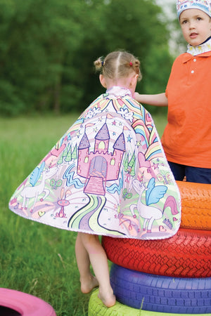 A child wearing the Great Pretenders Princess Color in Cape (for ages 4-7), featuring a colorful castle and unicorn design, stands on painted tires outdoors beside another child in an orange shirt.