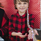 A child in a red and black plaid shirt with a Great Pretenders Storybook Braid Headband, Blonde in their hair, smiling at the camera against a red polka-dotted background.