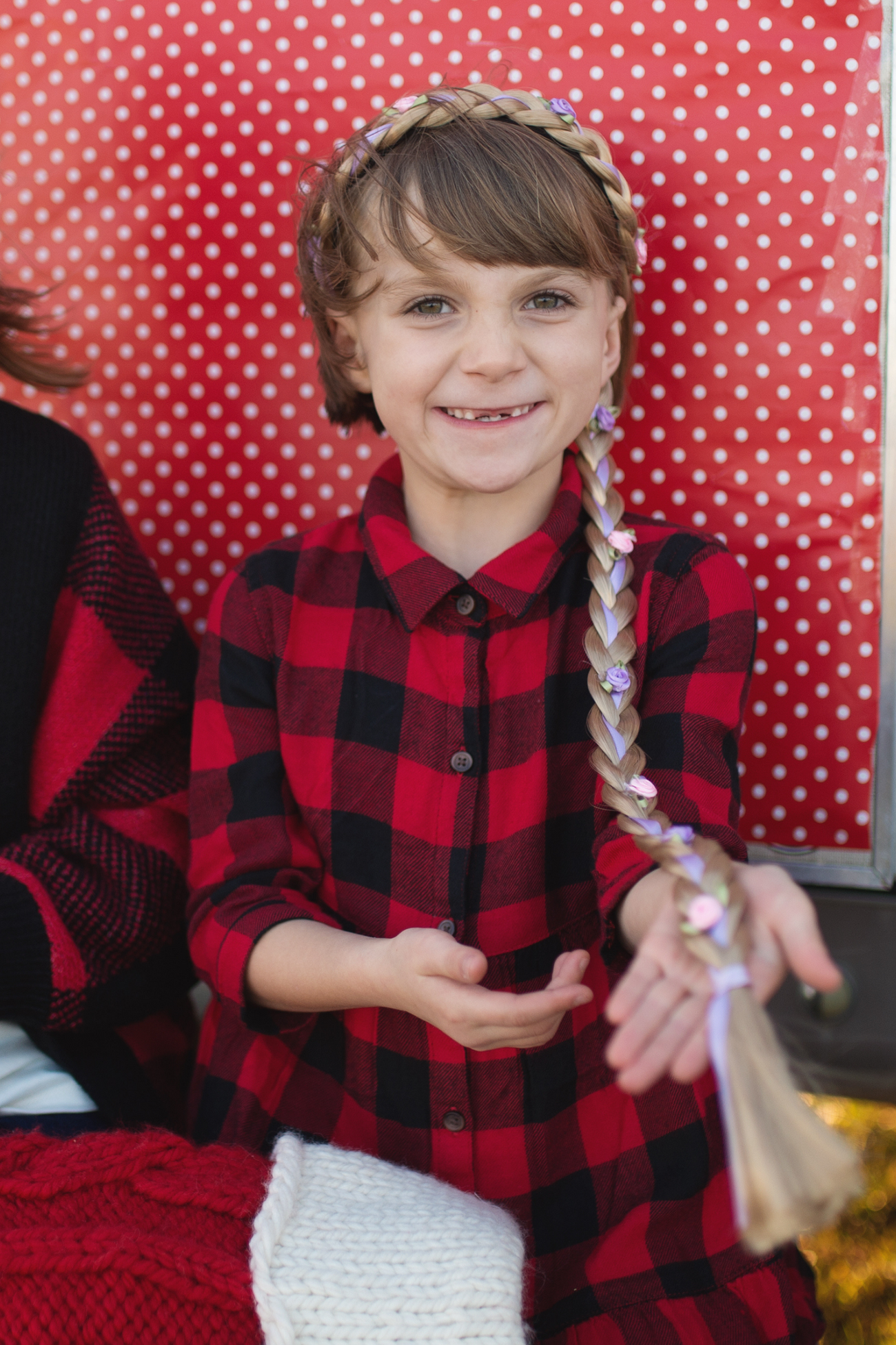 A child in a red and black plaid shirt with a Great Pretenders Storybook Braid Headband, Blonde in their hair, smiling at the camera against a red polka-dotted background.