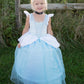 Young child with blonde hair in a bun wears the Great Pretenders Deluxe Cinderella Gown, made from premium satins and featuring a black choker, standing on grass with a wooden fence and plants behind.