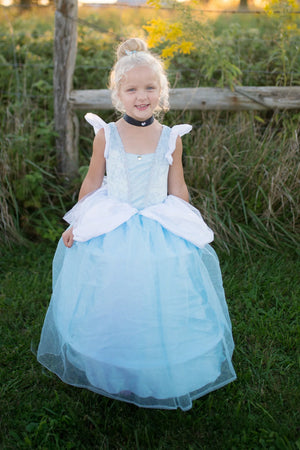 Young child with blonde hair in a bun wears the Great Pretenders Deluxe Cinderella Gown, made from premium satins and featuring a black choker, standing on grass with a wooden fence and plants behind.