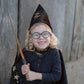 A smiling child with glasses wears a wizard hat and cape while holding the Great Pretenders Wizard Wand, ready to cast magical spells in front of a wooden wall.