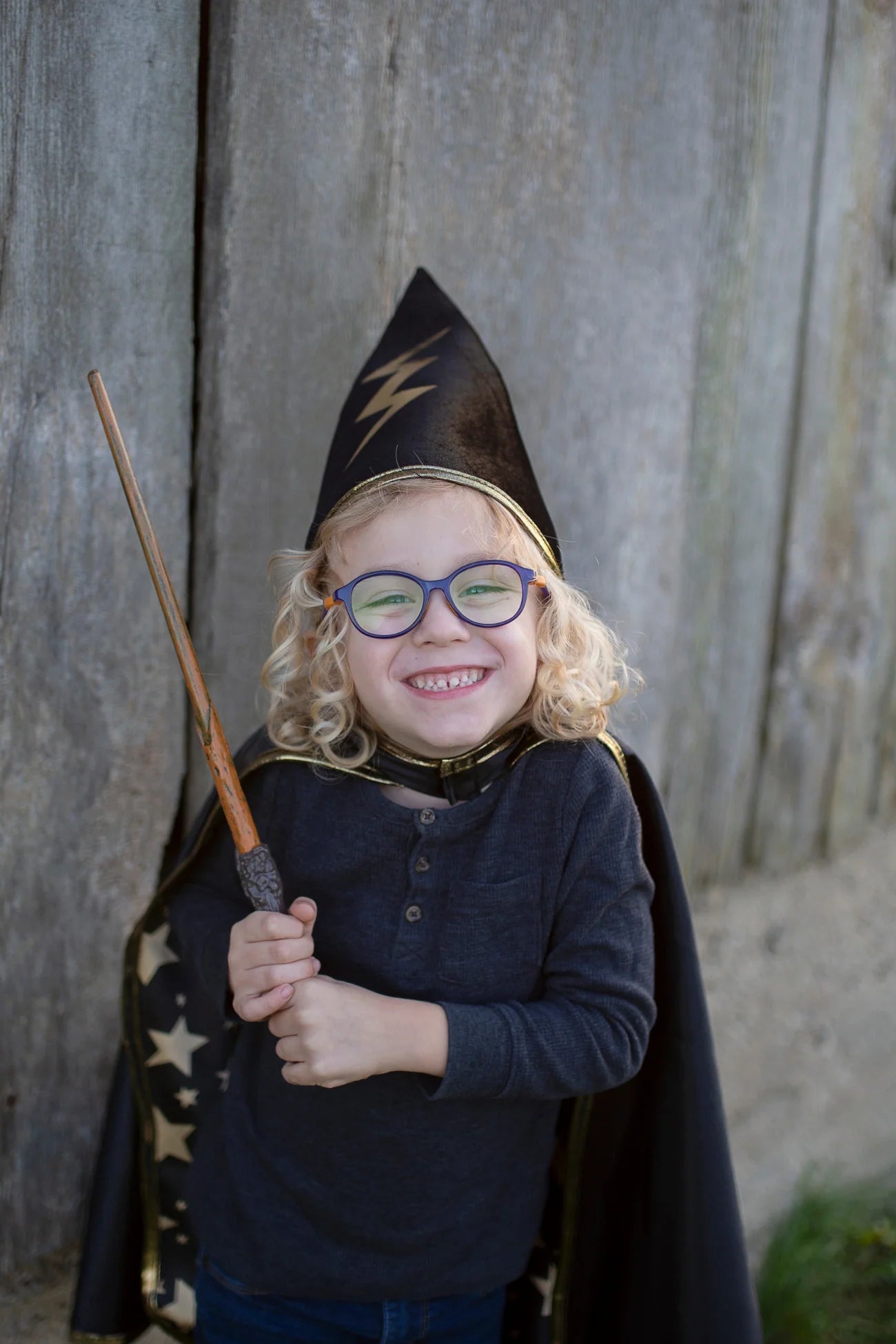 A smiling child with glasses wears a wizard hat and cape while holding the Great Pretenders Wizard Wand, ready to cast magical spells in front of a wooden wall.