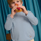 A young child wearing the Gingersnaps Boys’ Teo Jersey Tee, featuring dachshund embroidery, and beige pants, enjoys a hot dog with mustard while standing in front of a blue curtain.