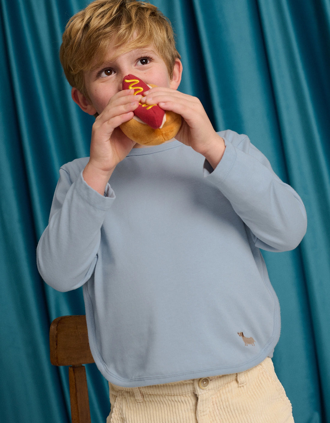 A young child wearing the Gingersnaps Boys’ Teo Jersey Tee, featuring dachshund embroidery, and beige pants, enjoys a hot dog with mustard while standing in front of a blue curtain.
