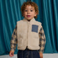 A young child with curly hair stands in front of teal curtains wearing a plaid shirt, dark jeans, and the Gingersnaps Baby Orazio Sherpa Vest featuring a blue pocket. This stylish outfit is perfect for layering babies on cooler days.