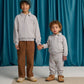 Two children stand on a beige rug holding hands, both wearing light gray French terry sweatshirts; one wears Gingersnaps Baby Boys’ Alessio French Terry Jogger Pants and a beanie, the other wears gray sweatpants. Blue curtains hang in the background.