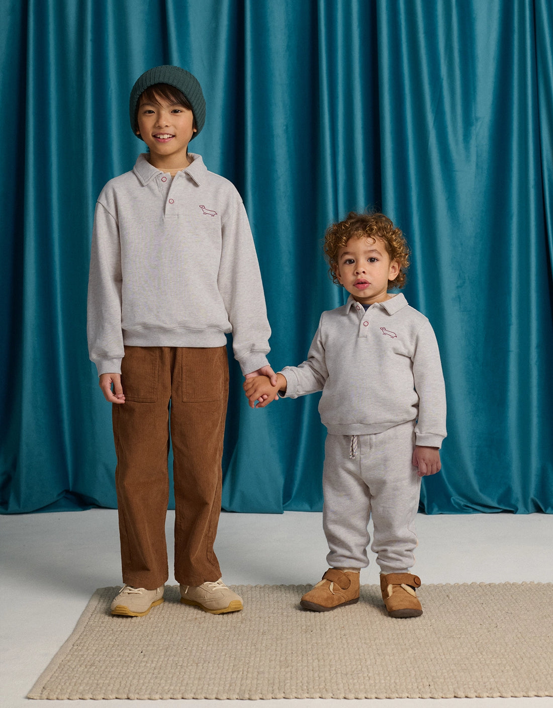 Two children stand on a beige rug holding hands, both wearing light gray French terry sweatshirts; one wears Gingersnaps Baby Boys’ Alessio French Terry Jogger Pants and a beanie, the other wears gray sweatpants. Blue curtains hang in the background.