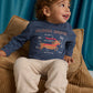 A toddler with curly hair sits on a tan corduroy chair, wearing the Gingersnaps Baby Boys’ Myron Graphic Tee featuring an embroidered outline dachshund illustration and beige pants, looking up and smiling.