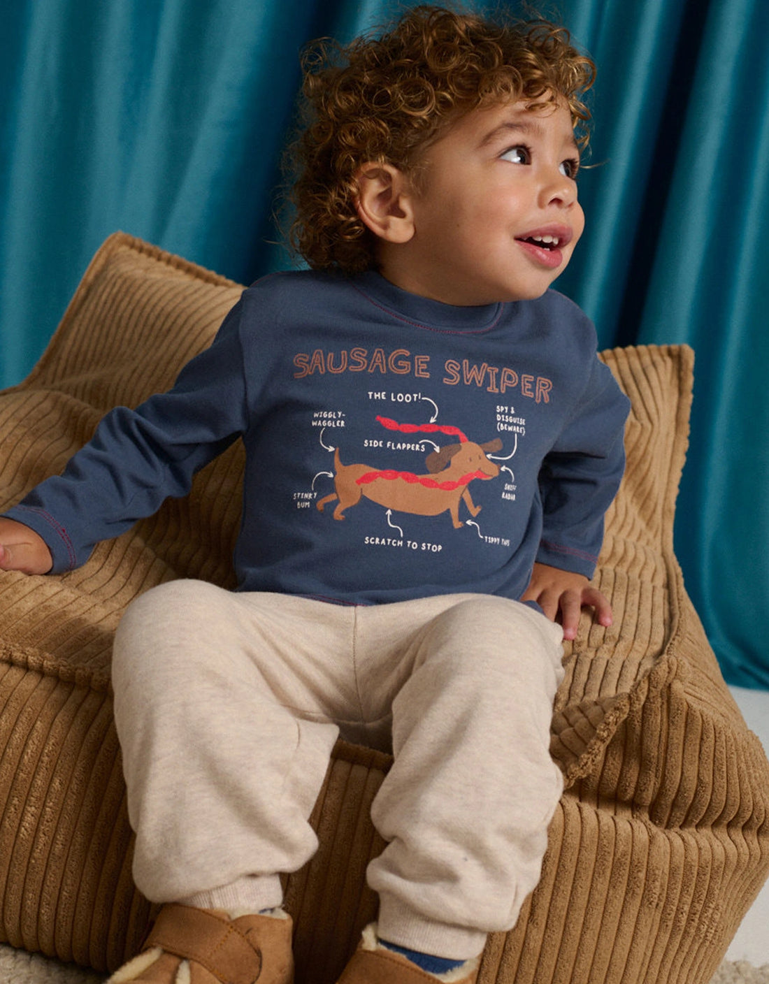 A toddler with curly hair sits on a tan corduroy chair, wearing the Gingersnaps Baby Boys’ Myron Graphic Tee featuring an embroidered outline dachshund illustration and beige pants, looking up and smiling.