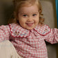 A toddler with light brown hair and a small ponytail smiles while sitting on a green chair, wearing the Gingersnaps Baby Girls’ Gracia Checkered Blouse and beige pants.