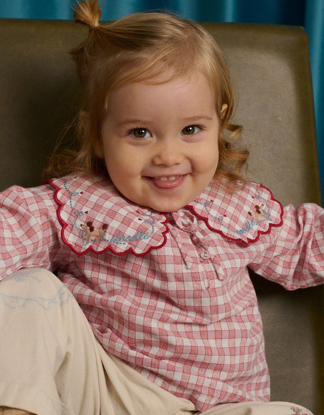 A toddler with light brown hair and a small ponytail smiles while sitting on a green chair, wearing the Gingersnaps Baby Girls’ Gracia Checkered Blouse and beige pants.