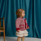 A toddler wears the Gingersnaps Baby Girls’ Lavinia Boxy Quilted Coat in red checkered fabric with bow-tie details, paired with a white dress and beige knee-high socks, standing indoors beside a wooden chair against a blue curtain backdrop.