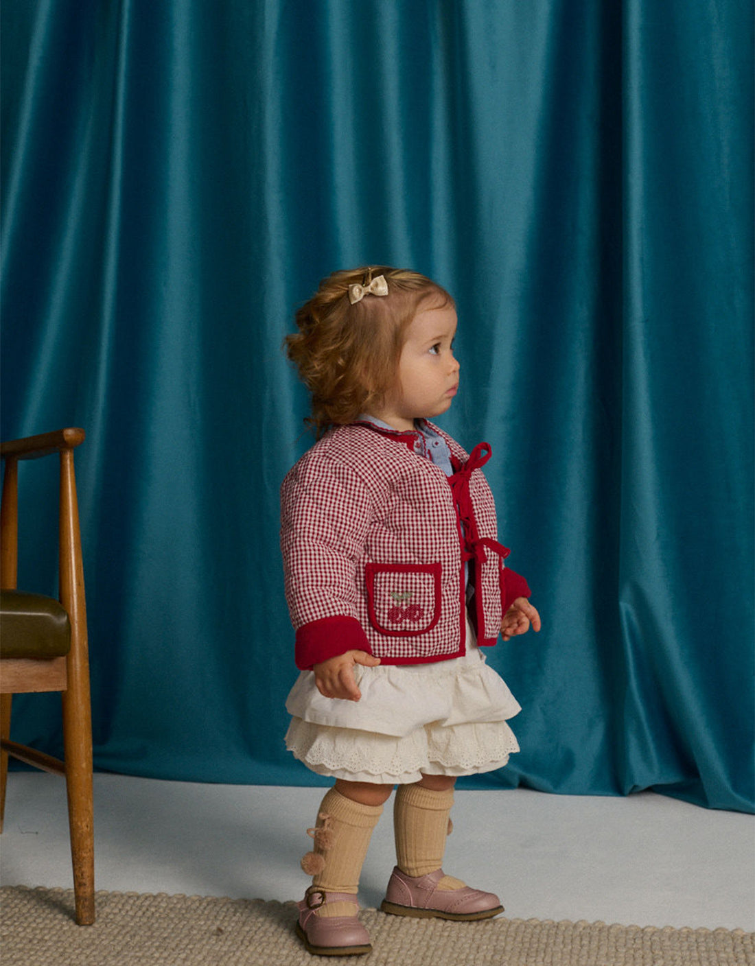 A toddler wears the Gingersnaps Baby Girls’ Lavinia Boxy Quilted Coat in red checkered fabric with bow-tie details, paired with a white dress and beige knee-high socks, standing indoors beside a wooden chair against a blue curtain backdrop.