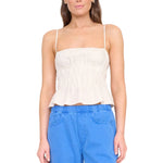 A woman wears the Staud Ayla Linen Tank, a sleeveless light beige cami with a smocked bodice and ruched peplum, paired with blue pants against a white background.