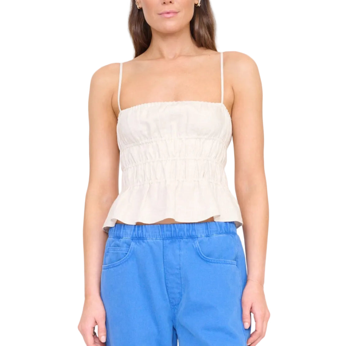 A woman wears the Staud Ayla Linen Tank, a sleeveless light beige cami with a smocked bodice and ruched peplum, paired with blue pants against a white background.