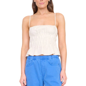 A woman wears the Staud Ayla Linen Tank, a sleeveless light beige cami with a smocked bodice and ruched peplum, paired with blue pants against a white background.