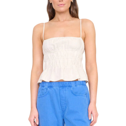 A woman wears the Staud Ayla Linen Tank, a sleeveless light beige cami with a smocked bodice and ruched peplum, paired with blue pants against a white background.