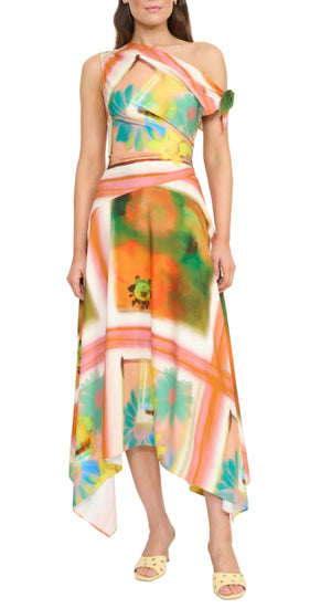 A woman wears the Staud Portia Dress, an asymmetrical, sleeveless dress with a colorful floral and abstract pattern and asymmetrical hem, paired with open-toe sandals.