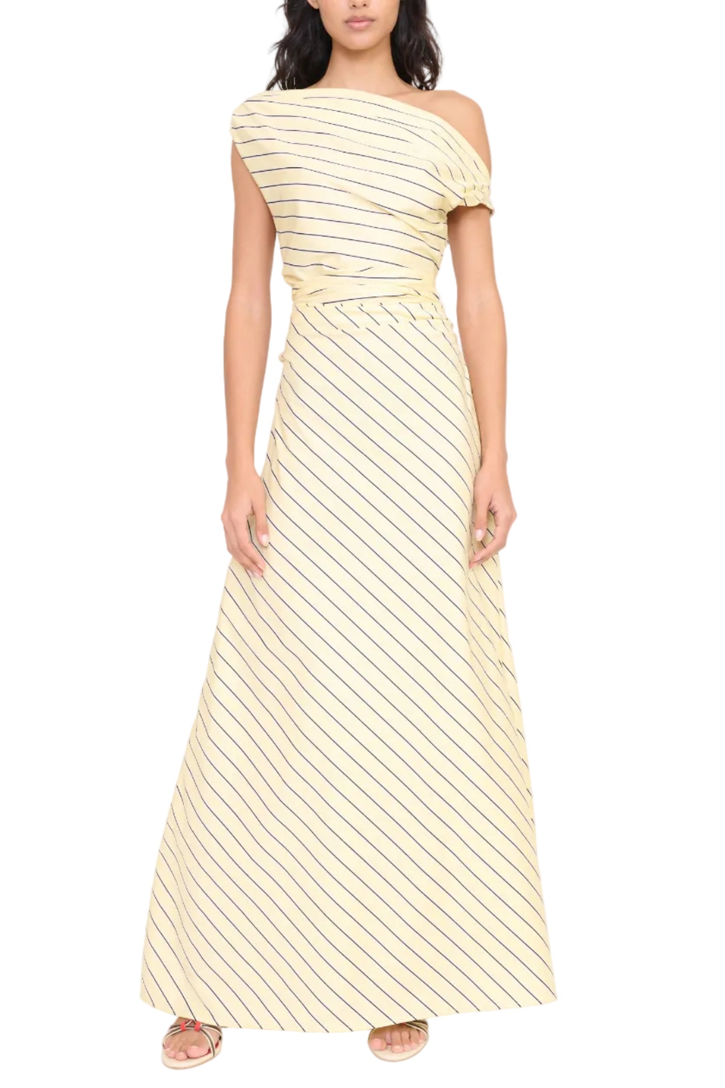 A woman stands against a white background wearing the Staud Phare Maxi Dress—an off-the-shoulder, floor-length yellow dress with diagonal black stripes—paired with flat sandals.
