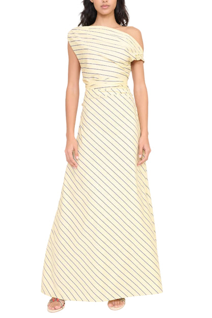 A woman stands against a white background wearing the Staud Phare Maxi Dress—an off-the-shoulder, floor-length yellow dress with diagonal black stripes—paired with flat sandals.