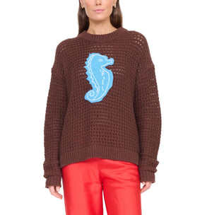 A person models the Staud Giada Seahorse Sweater, a brown drop shoulder style with a blue seahorse design, paired with red pants against a white background.