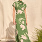A person stands outdoors in the Lela Rose Abstract Floral Crepe Draped Gown, featuring a straight-fit silhouette and short sleeves, posed before a neutral fabric backdrop.