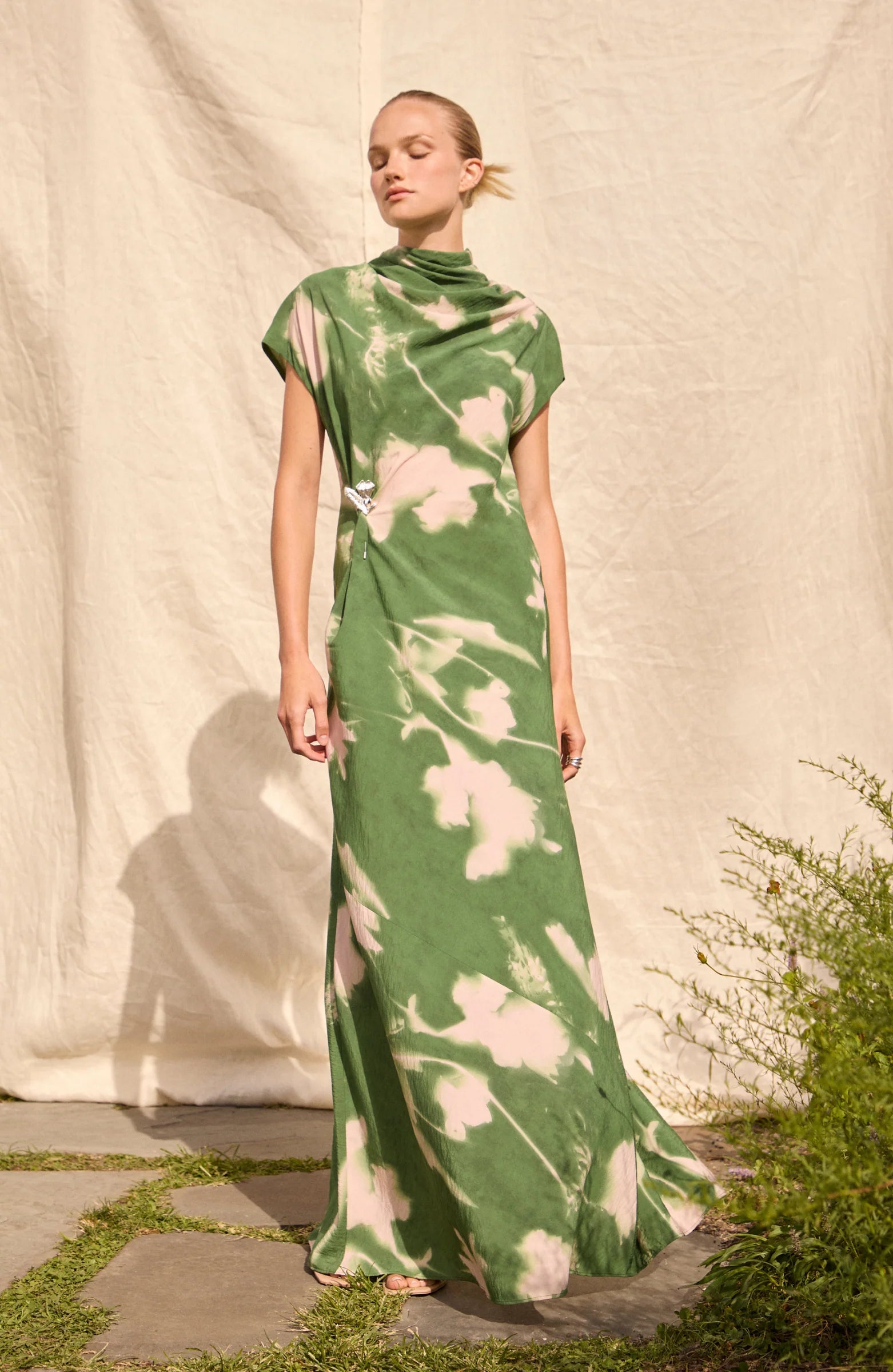 A person stands outdoors in the Lela Rose Abstract Floral Crepe Draped Gown, featuring a straight-fit silhouette and short sleeves, posed before a neutral fabric backdrop.