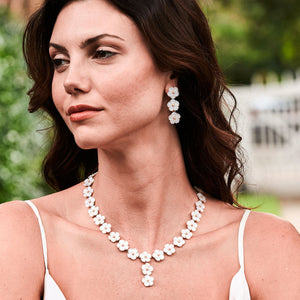 A woman outdoors wears the Juliska Capucine De Wulf Mermaid Garden Petite Riviere Convertible Necklace and matching earrings, her long dark hair flowing over a white top with greenery in the background.