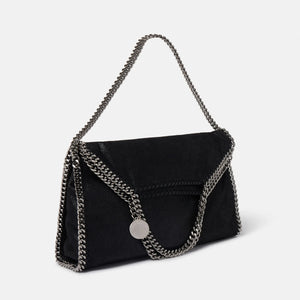 The Falabella Fold Over Tote by Stella McCartney is a black envelope-style vegan bag with chain edges and strap, plus a round metallic charm. Crafted from recycled plastic bottles, it's inspired by the iconic Falabella tote.