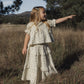 A young girl stands in tall grass, pointing to the right with trees and hills in the background, wearing the Rylee & Cru Girls’ Colette Top—a light-colored, patterned piece featuring smocked detailing.
