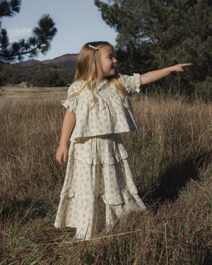 A young girl stands in tall grass, pointing to the right with trees and hills in the background, wearing the Rylee & Cru Girls’ Colette Top—a light-colored, patterned piece featuring smocked detailing.