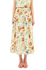 A person wearing the Ulla Johnson Theodora Skirt, an ankle-length floral piece with a gathered waist, paired with orange strappy sandals, stands against a white background.