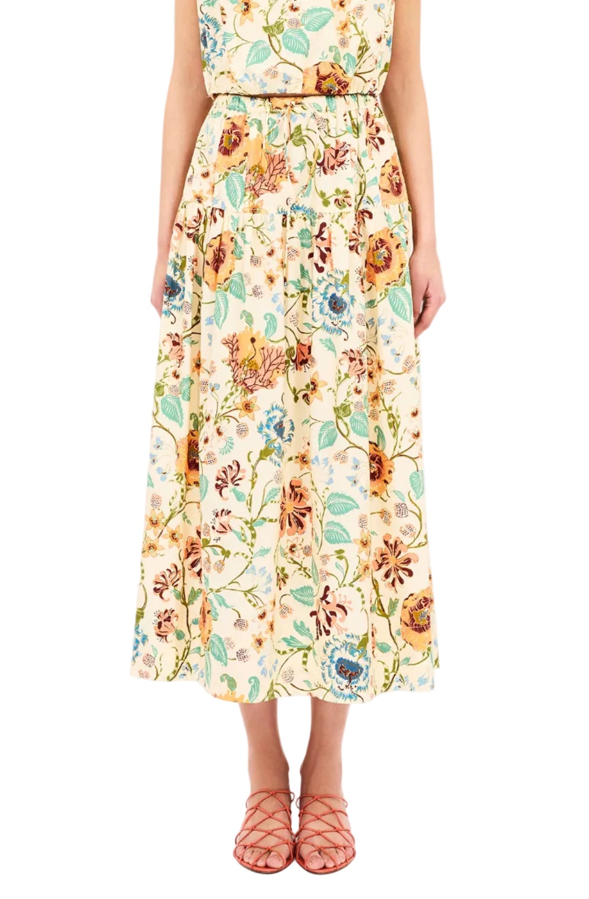 A person wearing the Ulla Johnson Theodora Skirt, an ankle-length floral piece with a gathered waist, paired with orange strappy sandals, stands against a white background.